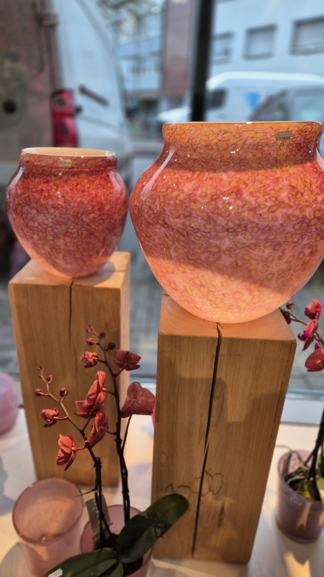 Zwei dekorative Glaskrüge in rosa und lila Tönen auf Holzständern, umgeben von kleinen Orchideenblüten.