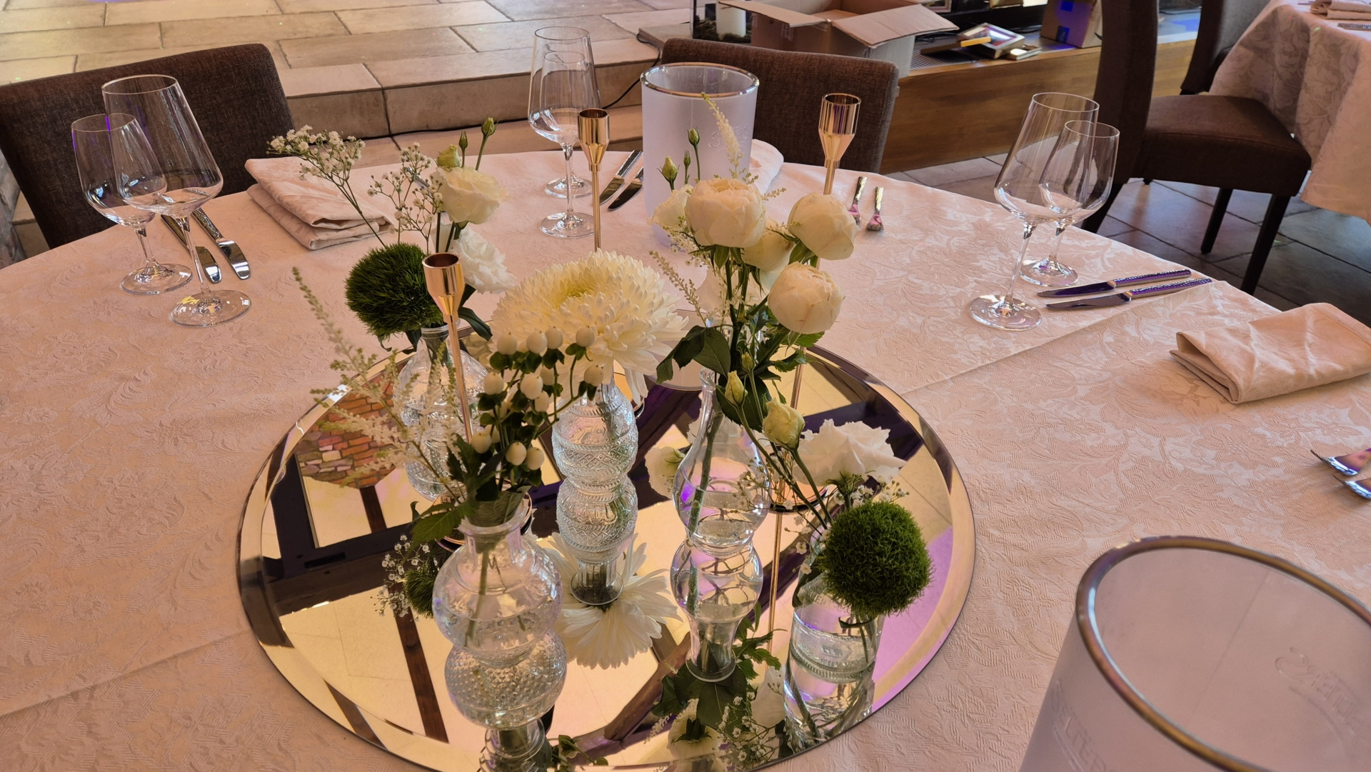 Tischdekoration mit weißen Blumen und grünen Akzenten auf einem runden Spiegel, umgeben von Gläsern und Besteck.